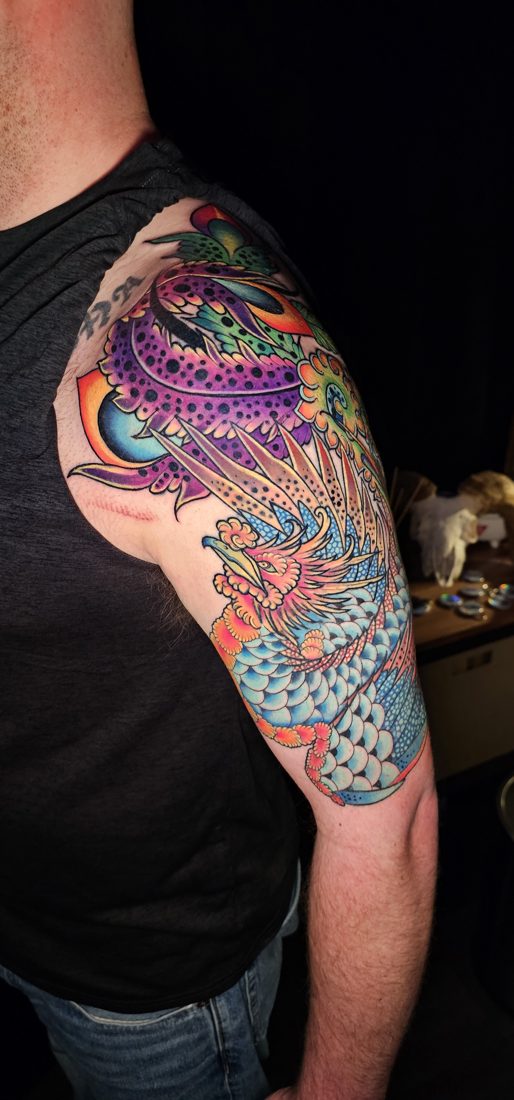 Ornate bird in saturated colour, tattoo across an upper arm and shoulder